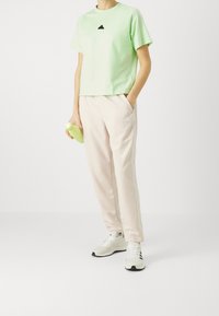 Grünes, kurzes T-Shirt mit schwarzem Logo, kombiniert mit hellrosa Jogginghosen. Die Person hält eine hellgrüne Wasserflasche. Weiße Sneakers sind zu sehen.