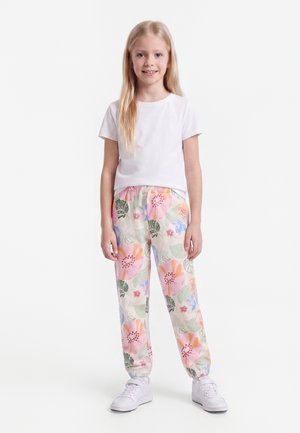 Νεαρό κορίτσι στέκεται, φορώντας ένα απλό λευκό μπλουζάκι, φλοράλ παντελόνι σε ροζ, πορτοκαλί, πράσινο και λευκά αθλητικά παπούτσια με φόντο λευκό.