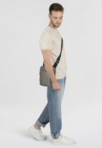 Borsa a tracolla in pelle grigia con tracolla nera regolabile, forma rettangolare, texture liscia, zip dettagliata, indossata da un modello in abbigliamento casual.