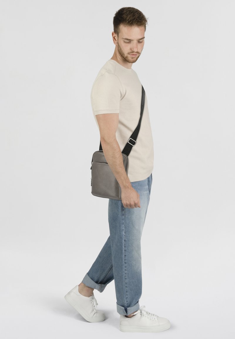 Borsa a tracolla in pelle grigia con tracolla nera regolabile, forma rettangolare, texture liscia, zip dettagliata, indossata da un modello in abbigliamento casual.
