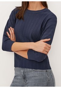 Femme portant un pull côtelé bleu marine à manches trois quarts et un jean en denim gris clair, les bras croisés sur la poitrine.
