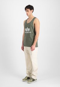 Tricou fără mâneci de culoare verde măslinie cu logo-ul alb "Alpha Industries", asortat cu pantaloni cargo crem și adidași verzi și gri.