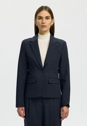 Vrouw met steil bruin haar die zilveren oorringen draagt, een witte coltrui en een donkerblauw jasje en broek, staand tegen een effen achtergrond.