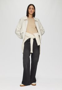 Helle, strukturierte Jacke mit zwei Fronttaschen, beige-weiß gestreiftes Hemd, schwarze weit geschnittene Jeans und beigefarbene Schuhe mit braunen Sohlen.