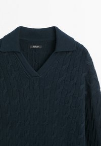 Marineblå kabelstrikket sweater med V-hals og opklappet krave, der har teksturerede snoede mønstre og et mærke ved halsudskæringen.