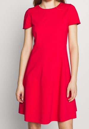 Femme portant une robe rouge vif à manches courtes, arrivant au genou, avec une taille cintrée et un décolleté rond, devant un fond clair.