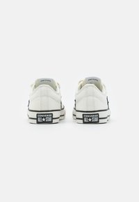 White canvas sneakers with black accents, featuring a low-top design, textured fabric, and Converse All Star branding on the heel.