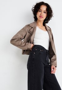 Svijetlosmeđa jakna od umjetnog brušenog antilopa s ovratnikom s prorezom i prednjim kopčanjem na kopče, nošena preko bijelog rebrastog topa i visokozračnih crnih traperica.