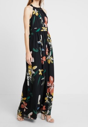 Femme portant une robe maxi noire sans manches avec de grandes fleurs multicolores imprimées et des sandales à talons blocs beiges à bout ouvert.
