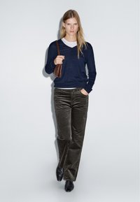 Maglione a v-neck blu navy, maglietta grigia sotto, pantaloni in corduroy oliva e scarpe nere; tiene una piccola borsa a mano marrone. Design semplice e pulito.