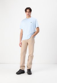 Tricou bleu deschis cu guler rotund și un mic logo verde, purtat cu pantaloni bej cu croială dreaptă și adidași negri cu textură.