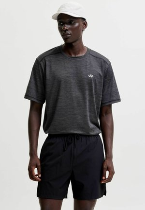 Mand iført en mørkegrå kortærmet skjorte, sorte shorts og en hvid kasket, stående mod en ensfarvet lys baggrund.