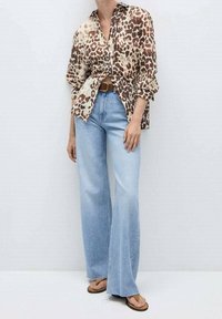 Femme portant un chemisier imprimé léopard marron et beige, un jean clair taille haute évasé, une ceinture marron et des sandales marron, debout contre un mur blanc.