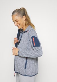 Sweaterjack in een lichtblauw en marineblauw visgraatpatroon. Kenmerkt zich door een ritszak op de linkerarm, contrasterende manchetten en een ritsluiting.
