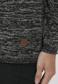 Black and gray, knitted fabric pullover with a textured pattern. Features a brown rectangular leather tag with embossed logo on the hem.