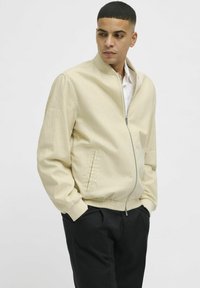Veste bomber beige avec un col montant, fermeture éclair sur le devant, poches latérales et poignets élastiques. Portée sur une chemise légère, associée à un pantalon foncé.