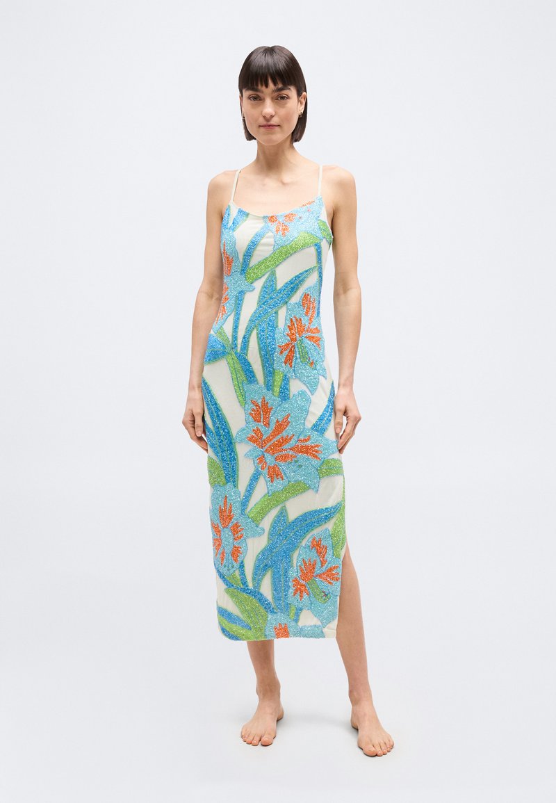 Γυναίκα που στέκεται ξυπόλητη φορώντας μίντι αμ άνικο φόρεμα με μπλε, πράσινα και πορτοκαλί λουλουδένια σχέδια σε λευκό φόντο.