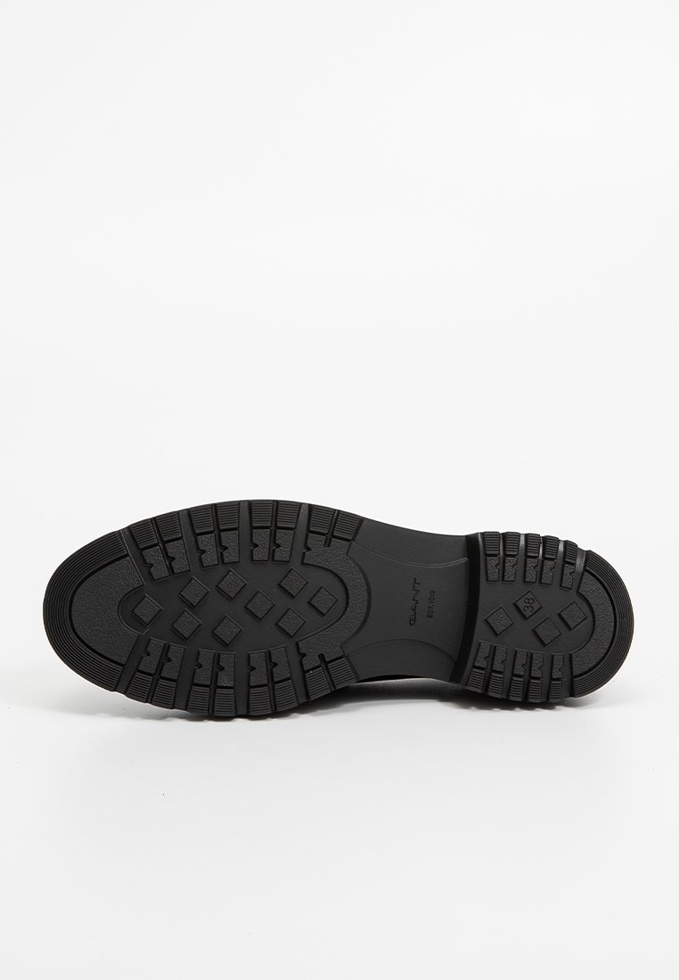 Schwarze Gummisohle mit strukturierten Traktionsmustern. Enthält geometrische Formen und das Markenlogo; auf einem weißen Hintergrund präsentiert.