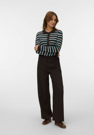 Femme aux cheveux bruns portant un haut rayé à manches longues, un pantalon large sombre et des chaussures marron, debout, les bras croisés, sur fond blanc.