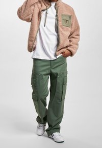 Polarowy sweter w jasnoróżowym kolorze z zieloną naszywaną kieszenią, połączony z zielonymi spodniami cargo z kieszeniami bocznymi i zatrzaskowymi detalami. Białe sneakersy.