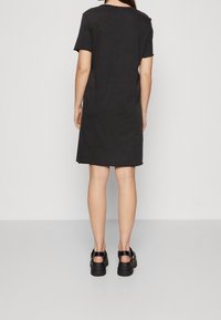 Mustan lyhythihaisen mekon suora leikkaus, joka on valmistettu pehmeästä materiaalista. Mekossa on pyöreä pääntie ja sivujen halkiot, yhdistettynä paksupohjaisiin mustiin kenkiin.