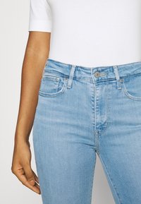 Persona vestida con jeans de mezclilla azul claro y una camisa de manga corta blanca, con la mano izquierda relajada al lado de la cadera.