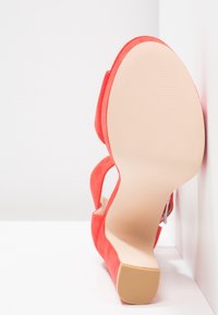 Korallinväriset mokkasaappaat, joissa on avonainen takaosa, kapea hihna ja sileä beige kumipohja. Pohjassa on kuvioitua pintaa.