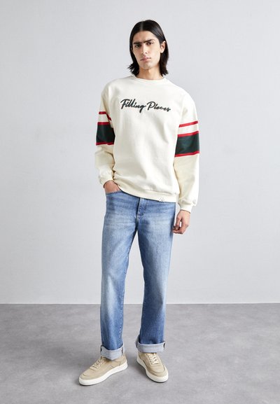 Cream sweatshirt with "Filling Pieces" logo, red and green stripes on sleeves, paired with light blue jeans and beige sneakers.