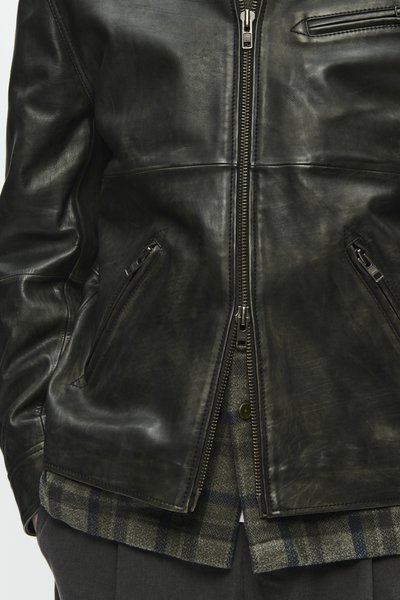 Black leather jacket featuring a zip closure and notable texture, with angled side pockets and a zipped chest pocket. Layered over a plaid shirt.