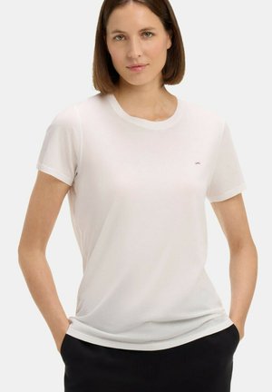 Femme aux cheveux courts bruns portant un t-shirt blanc uni à manches courtes et un pantalon noir, debout les mains dans les poches.