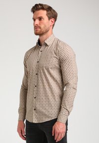 Camisa de manga larga abotonada en beige con un diseño de patrón geométrico. Presenta un cuello extendido y botones oscuros. Se lleva con pantalones negros.