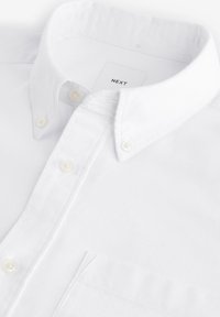 Camicia bianca con colletto classico, realizzata in tessuto liscio. Presenta piccoli bottoni bianchi e una singola tasca sul petto.