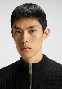 Giovane uomo con capelli neri corti che indossa un maglione zip-up a coste nere con colletto alto su uno sfondo chiaro e semplice.