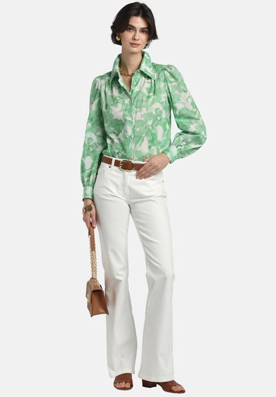 Sieviete, kura valkā zaļu ziedu raksta blūzi ar garām piedurknēm, baltas augsta vidukļa bikses, brūnu jostu, brūnas sandales un tur brūnu somiņu ar zelta ķēdi.