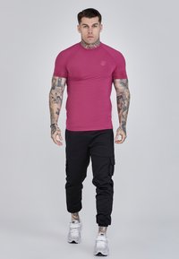 Īsām piedurkņu pieguļošs krekls magentas krāsā ar teksturētu audumu. Apvienots ar melno cargo biksēm ar sānu kabatām, valkāts ar baltām sporta apaviem.