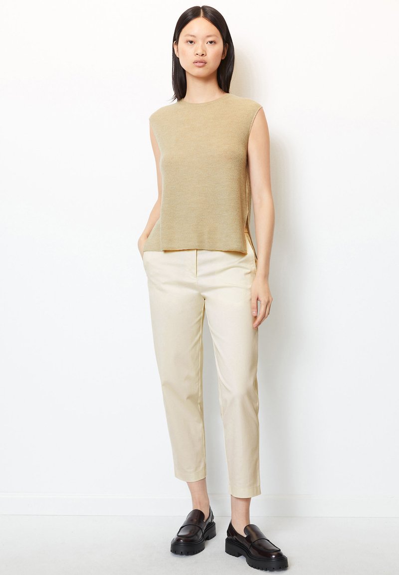 Beige, ärmelloses Stricktop mit lockerer Passform; kombiniert mit cremefarbenen, schmal zulaufenden Hosen; schwarze klobige Slipper runden das Outfit ab.