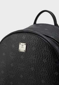 Mochila de cuero negro texturizado con logotipos MCM en relieve y una placa de metal plateada en el bolsillo frontal, que cuenta con un asa superior y cierre de cremallera.