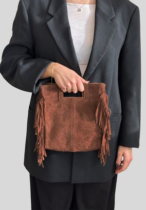 Borsa a mano in camoscio marrone con dettagli in frange, forma rettangolare e manico superiore. Tenuta da una persona che indossa un blazer nero e una maglia bianca.
