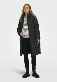 Abrigo largo negro acolchado con un cuello alto, combinado con una blusa de rayas blanca y negra y pantalones negros. Textura suave con cierre de cremallera.