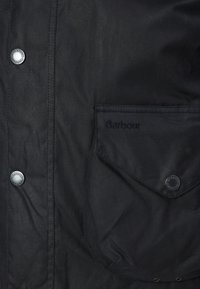 Fekete viaszolt dzseki gombos záródással, látható varrással és elülső zsebbel, amelyen "Barbour" feliratozás található. Tartós és texturált anyag.