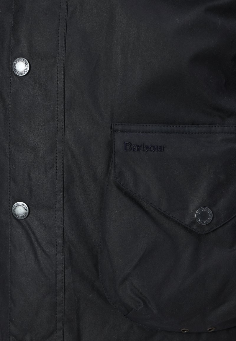 Schwarze gewachste Jacke mit Knopfverschluss, sichtbaren Nähten und einer Vorderentasche mit eingeprägtem "Barbour". Robuster und strukturierter Stoff.