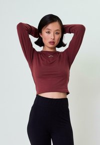 Kastanjetop murrus värviga pika varrukaga särk ümmarguse kaelusega, millel on rinnal väike logo, koos kõrge vöökohaga mustade pükstega.