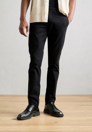 Homme portant un pantalon noir slim, des chaussures noires à lacets, une chemise beige à manches courtes, debout sur un sol en bois clair devant un fond gris.