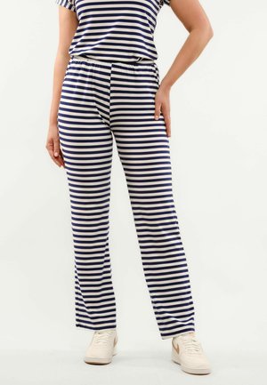 Osoba nosi tmavomodré a biele horizontálne pruhované nohavice a zodpovedajúci top, stojí s jednou rukou na boku a bielymi teniskami.