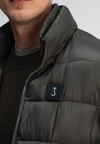 Dunkelgraue gepolsterte Jacke mit hohem Kragen, ausgestattet mit einem schwarzen Logopatch und subtiler Textur auf dem Stoff. Reißverschlussdetails sind sichtbar.