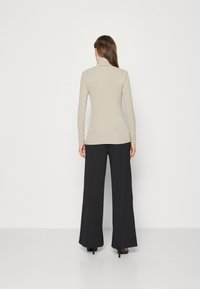 Donna di spalle che indossa un maglione a collo alto beige, pantaloni neri a gamba larga e sandali neri con tacco su uno sfondo bianco semplice.