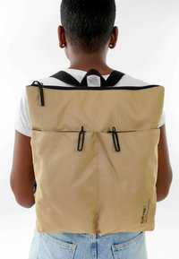 Mochila de tela beige con cremallera y cierres negros, que cuenta con dos bolsillos frontales; diseño simple con correas ajustables negras.