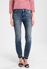 Hubené džíny z tmavě modrého denimového materiálu s mírným vyblednutím, s pěti kapsami a standardním zapínáním na knoflík. Nalezeno s otevřenými černými podpatky.