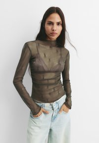 Langærmet, gennemsigtig brun mesh top med h�øj hals, der har et subtilt blomstermønster og er lagt over en mørk bh. Sættes sammen med lyseblå jeans.