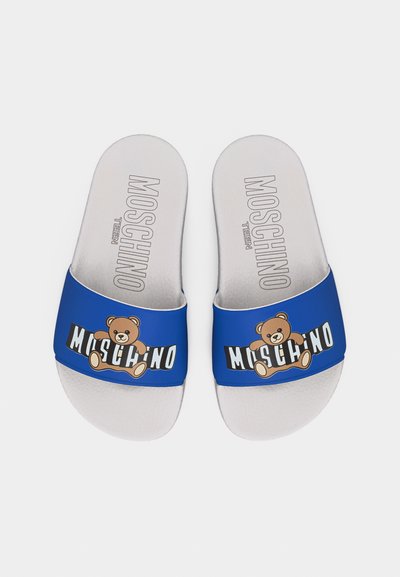 Λευκές σανδάλες με λουράκι μπλε, με καφέ αρκουδάκι και κείμενο «Moschino», με την επιγραφή «Moschino Teen» στα πέλματα.